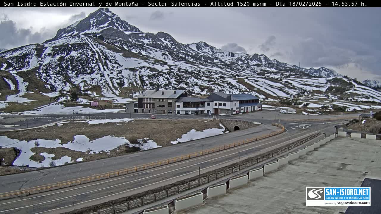 A snow-dusted mountainside overlooks a parking lot and road curving around a building complex under an overcast sky.