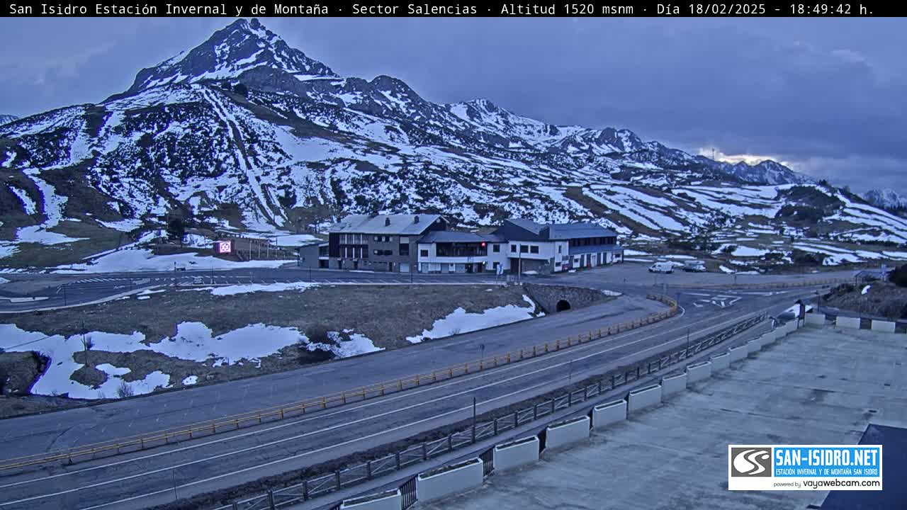 A mountain range partially covered in snow is seen under an overcast sky, with a road and buildings situated in the foreground.
