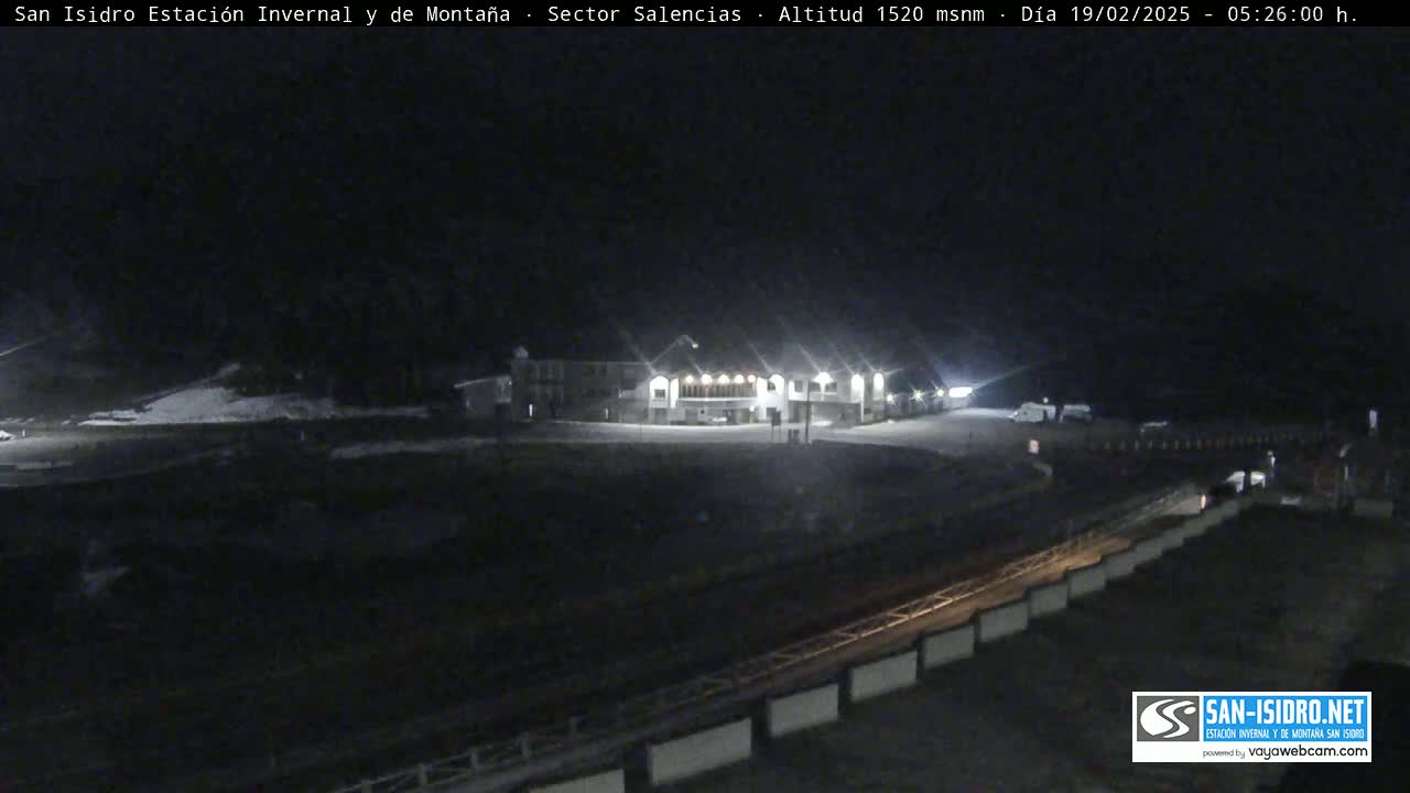 A nighttime view of a snow-covered parking lot and a lit building at a mountain resort under a dark sky.