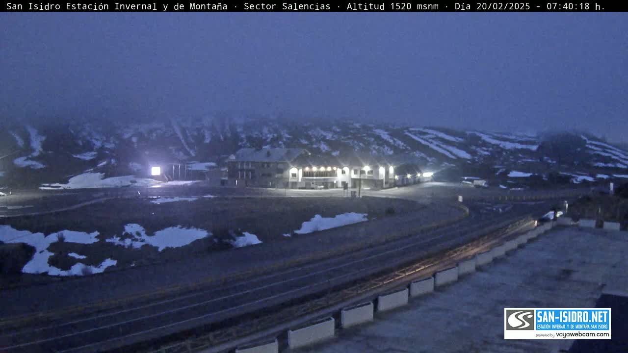 A dimly lit mountain lodge sits in a snow-dusted parking lot, under a dark, hazy sky.