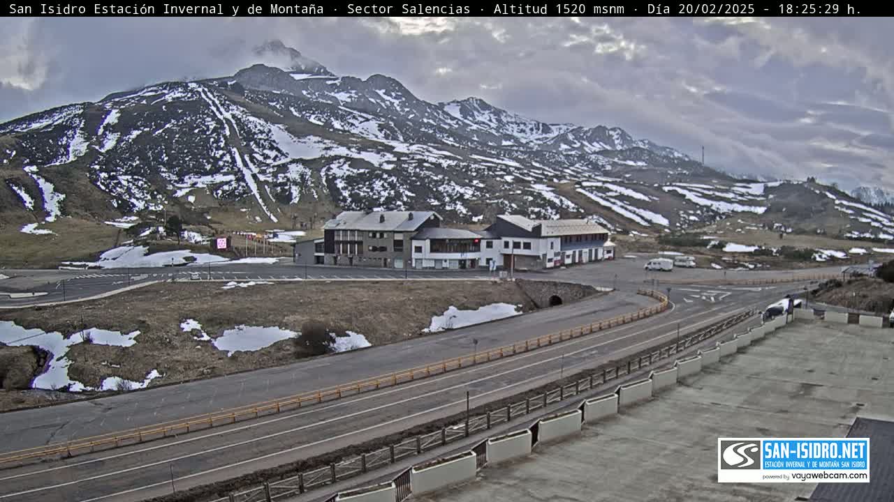 A mountain road winds past a complex of buildings near a snow-dusted mountain under a cloudy sky.