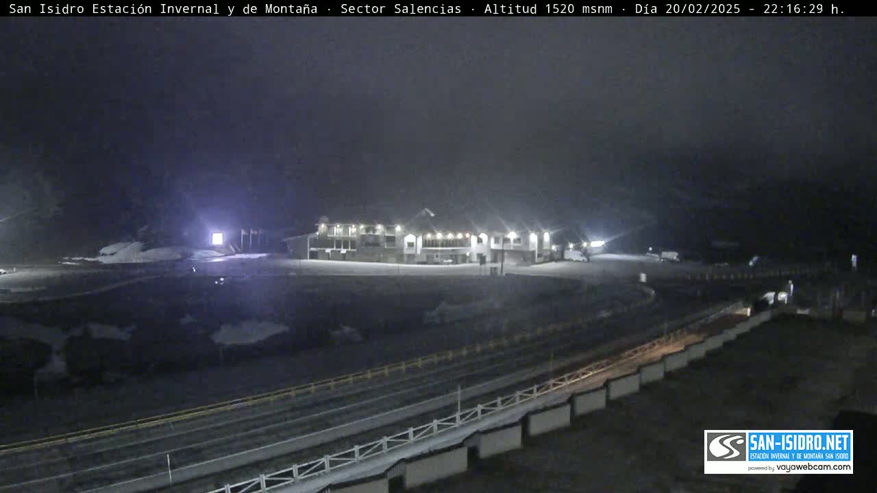 A snow-covered parking lot and roadway are illuminated at night in front of a building, under a dark, overcast sky.
