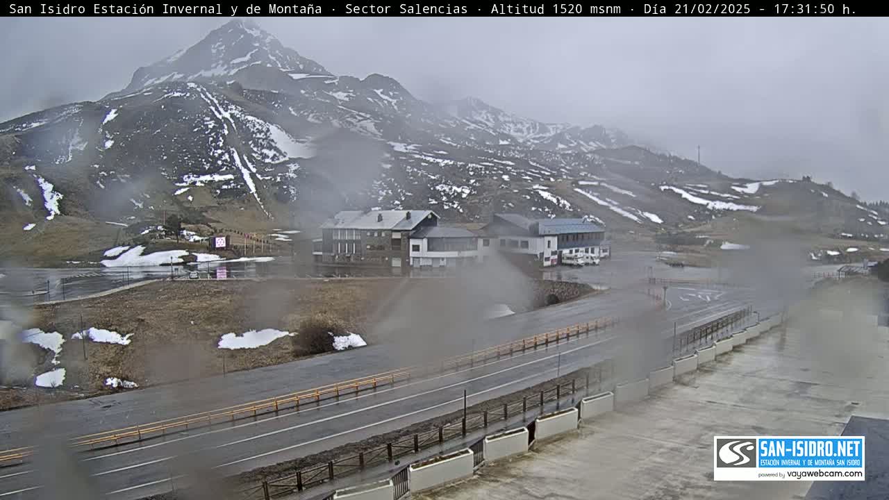A mountain range with patchy snow is visible in the distance under an overcast sky, with a road and several buildings in the foreground, all appearing wet from rain or mist.
