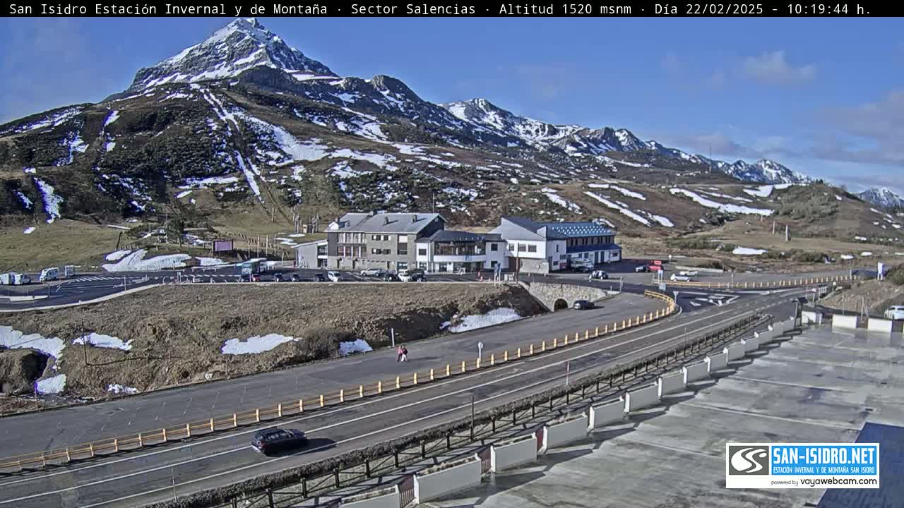 A mountain resort with a parking lot and buildings sits below a snow-dusted mountain range under a mostly sunny sky.