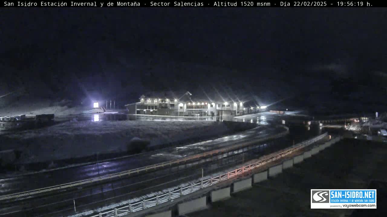 A nighttime view of a snow-covered roadway curving past a lit building, under a dark sky.