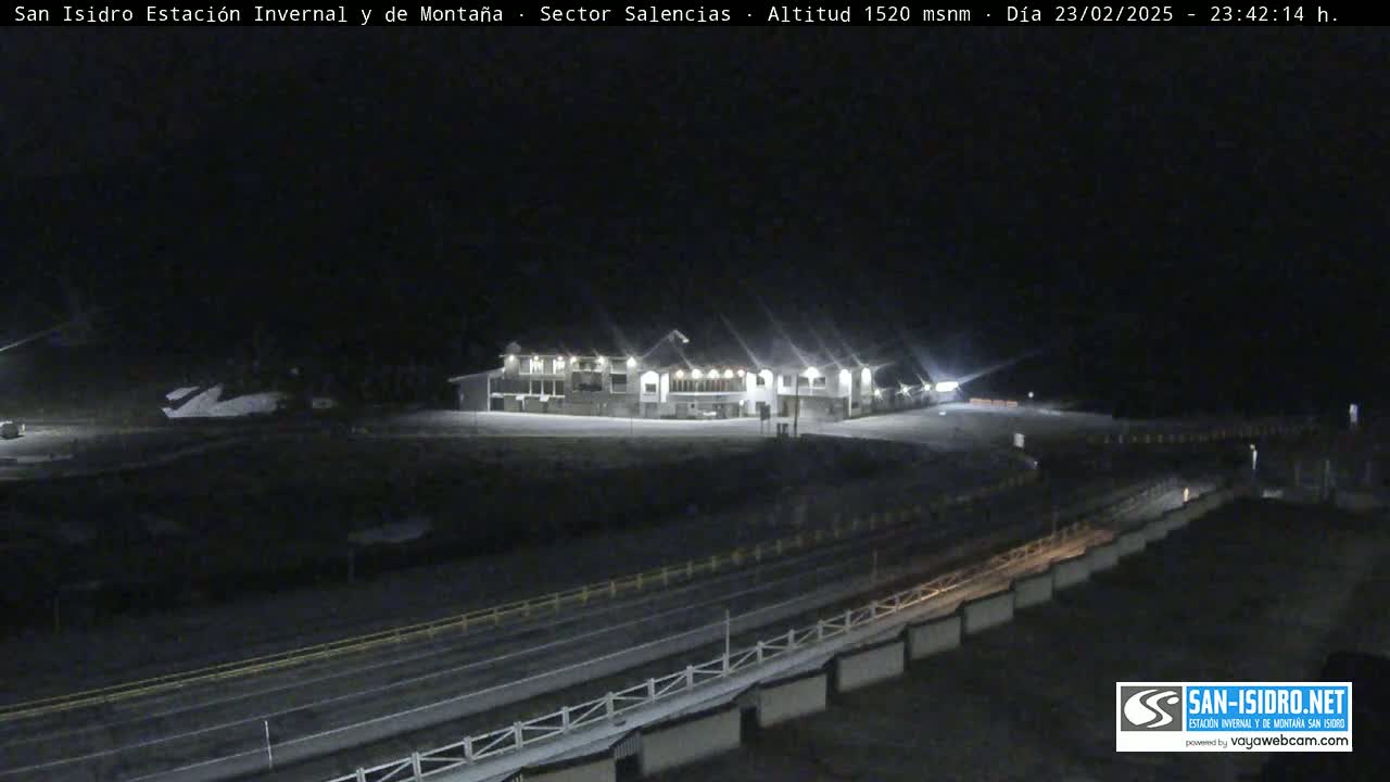 A long, low building is brightly lit at night in a snowy area, with a road and barrier in the foreground.