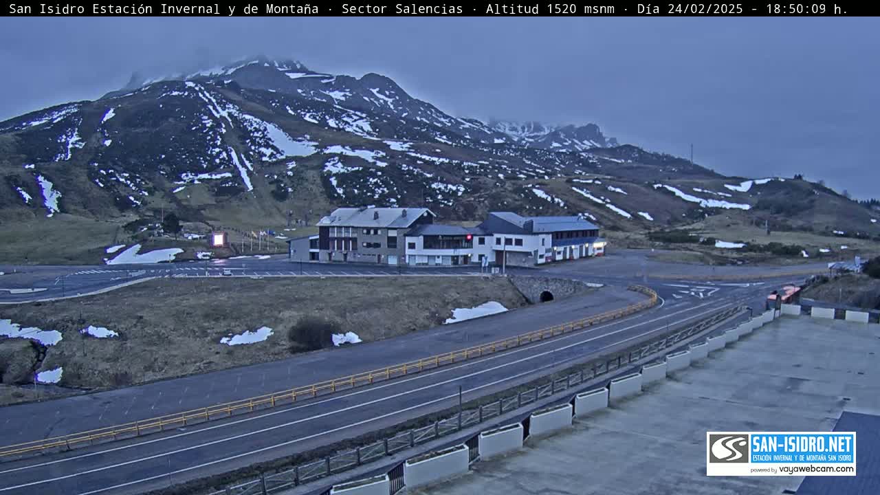 A mountain road winds past a complex of buildings, with snow-dusted mountains in the background under a twilight sky.