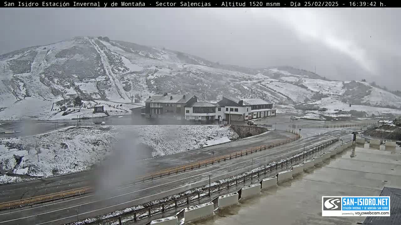 A snow-covered mountain slope with ski runs, a cluster of buildings, and roads are visible in hazy, snowy weather.