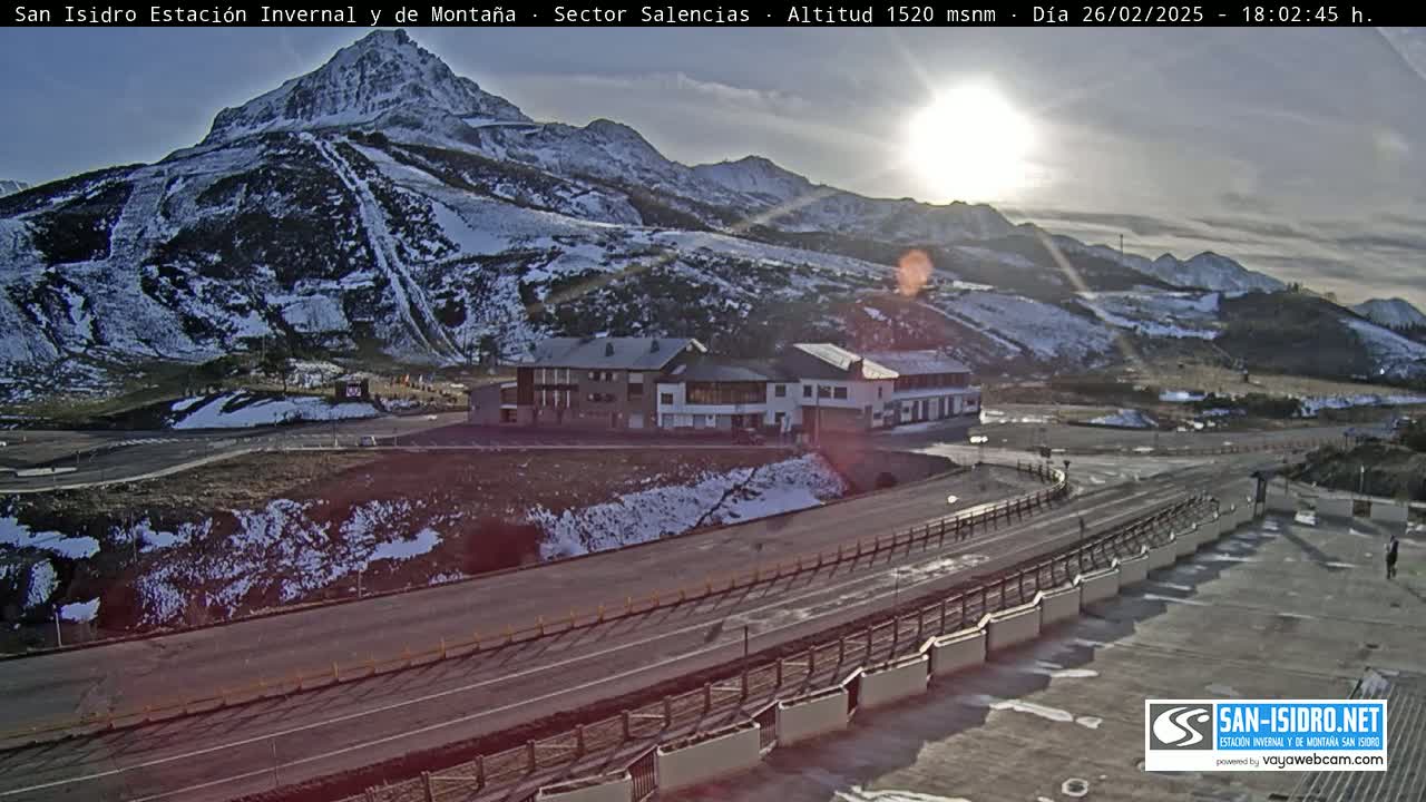A sunny, snow-dusted mountainside overlooks a road and building complex, with patches of snow visible on the ground.