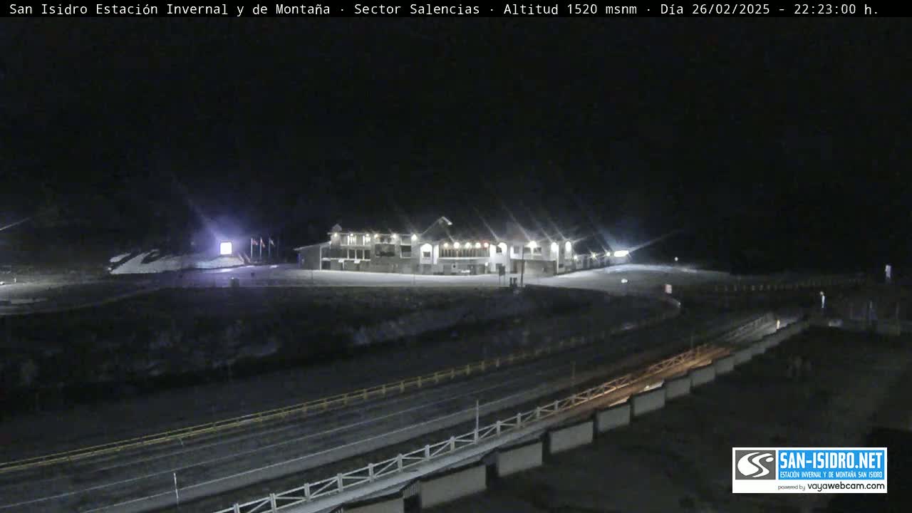 A large building is illuminated at night, with a snowy area in the foreground and a road below.