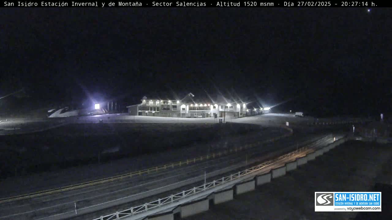 A long, low building is illuminated at night in a snowy area, with a road and guardrail visible in the foreground.