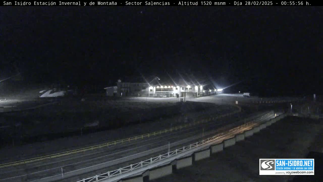 A brightly lit building sits in a snow-covered area at night, with a road and guardrail visible in the foreground.