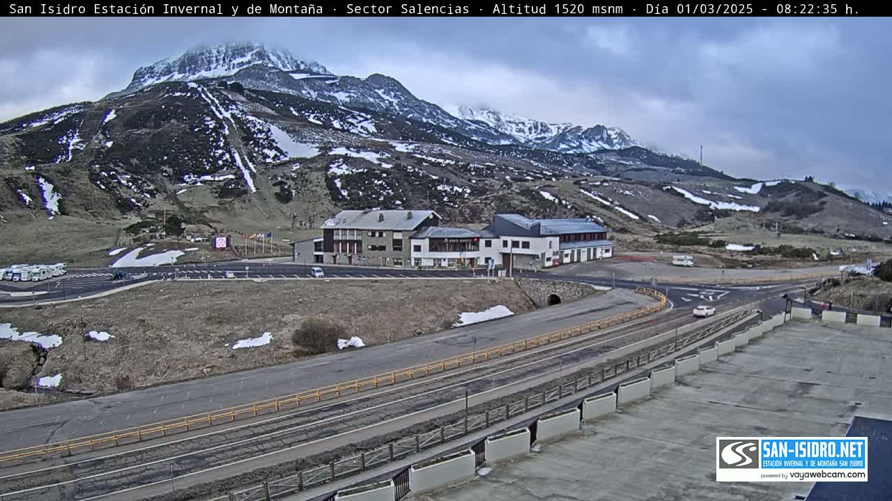 A mountain resort with a parking lot and several buildings sits below a snow-dusted mountain range under an overcast sky.