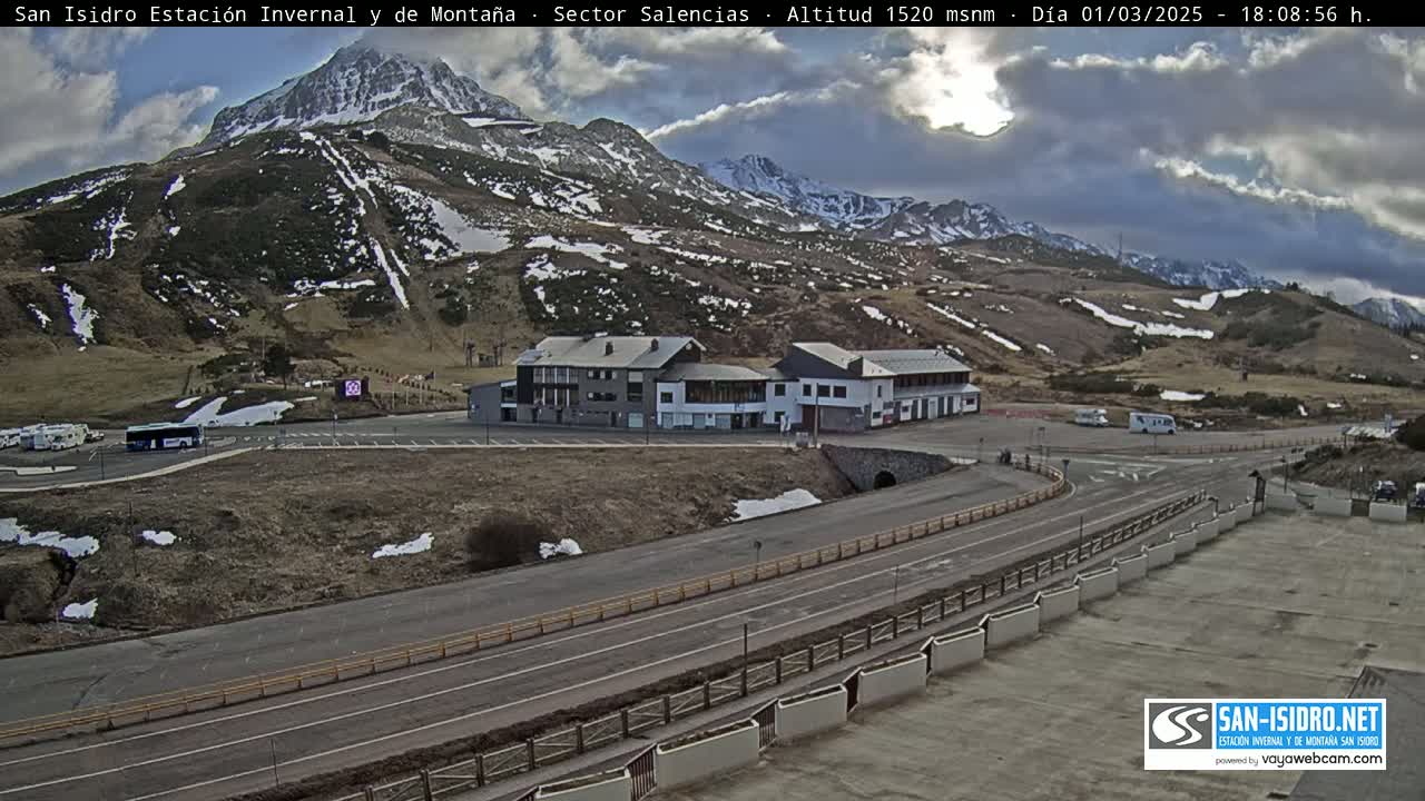 A mountainside resort with a parking lot and buildings, partially covered in snow, under a partly cloudy sky.