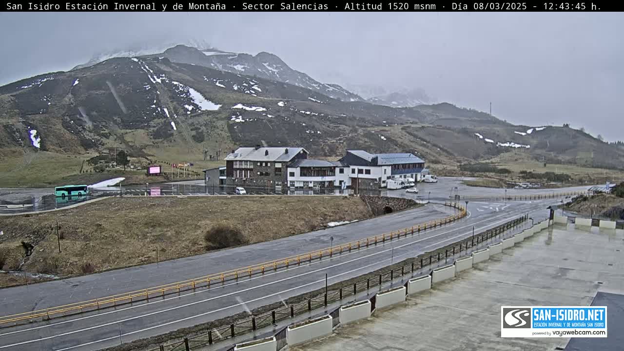 A mountain road winds past a parking lot and several buildings, all situated against a backdrop of overcast mountains speckled with patches of snow.