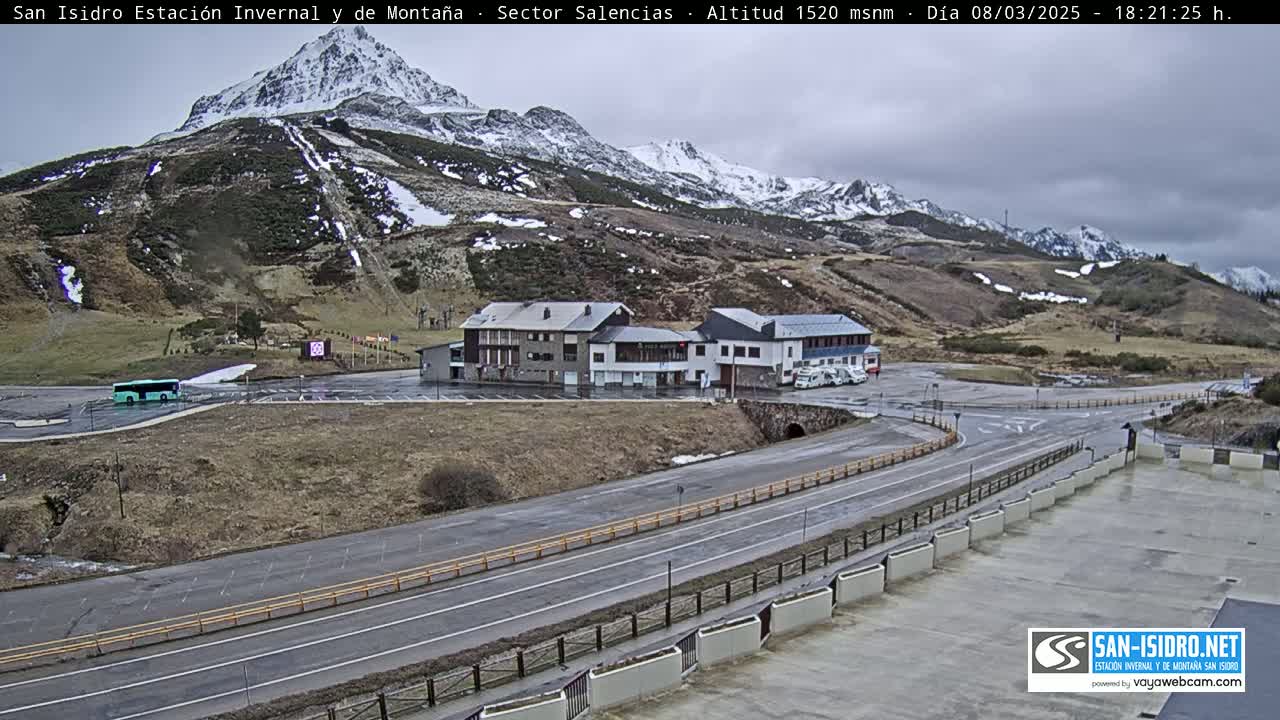 A mountainside parking lot with two buildings and a road is visible under an overcast sky, with snow on the distant mountains.