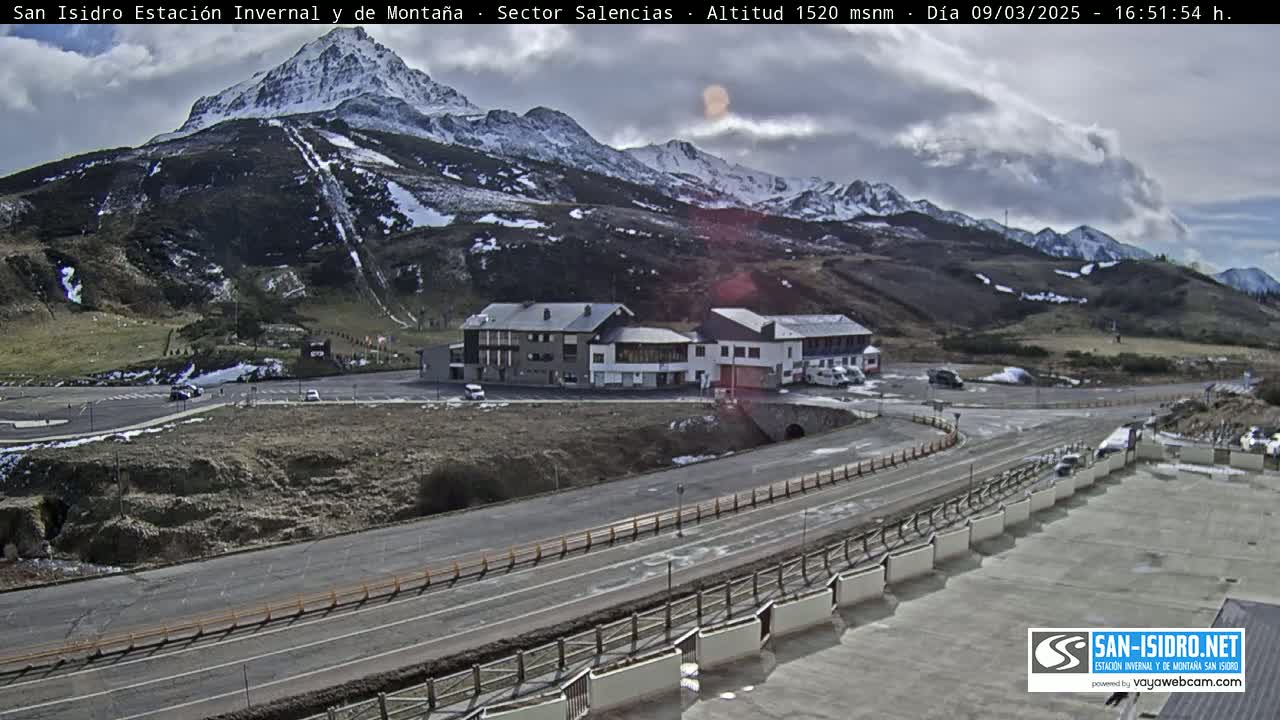 A mountain resort with a parking lot and buildings sits below a snow-capped mountain range under a partly cloudy sky.