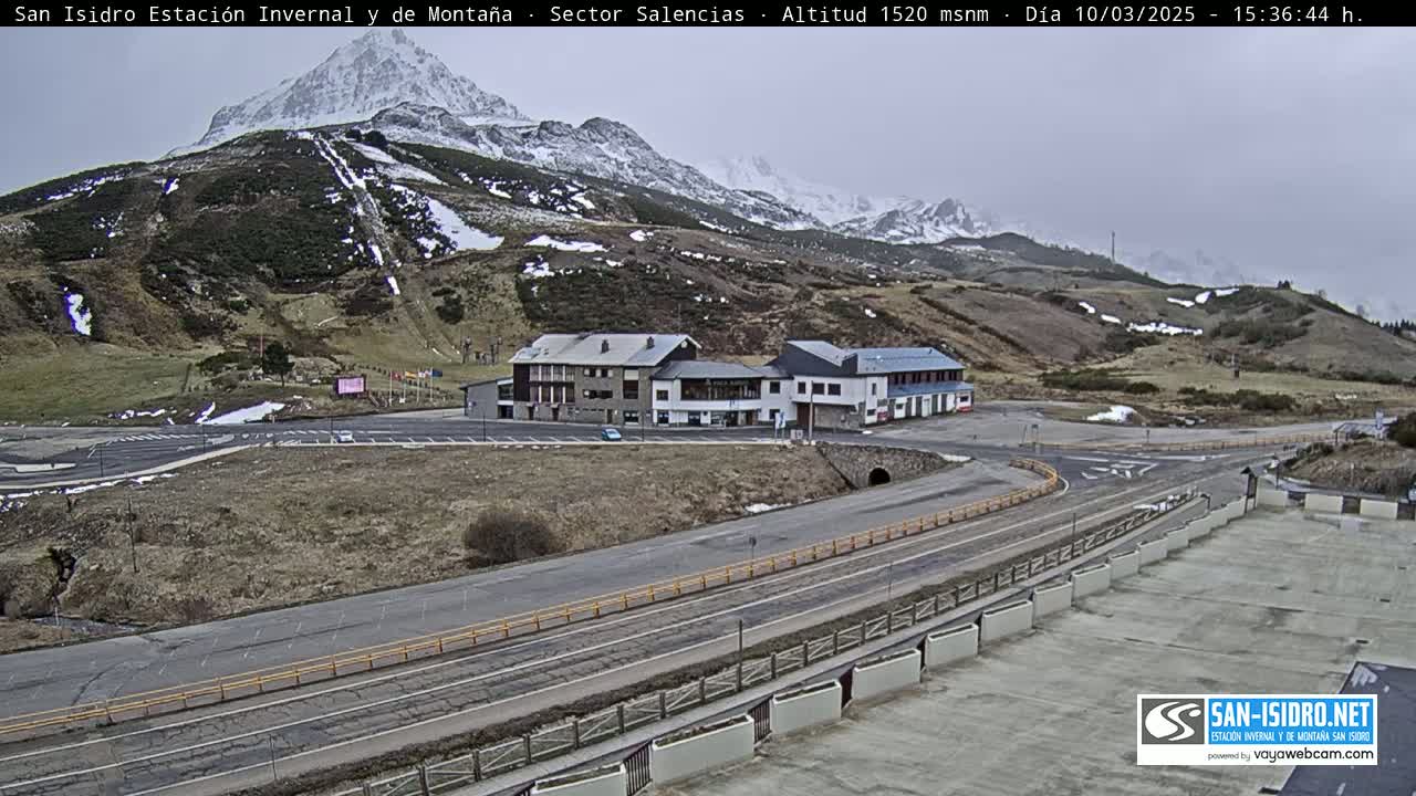 A mountain resort area with snow-capped mountains in the background, under an overcast sky, features buildings, roads, and parking areas.