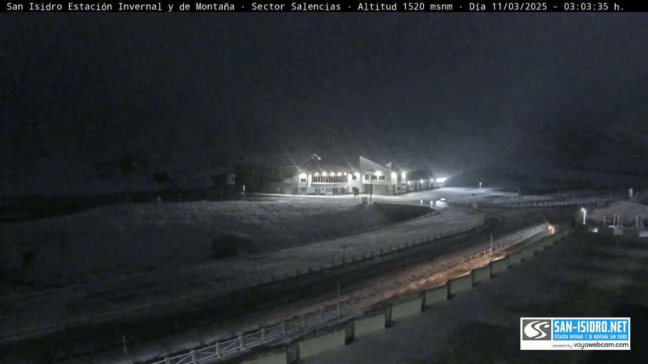 A snow-covered mountain road curves past a lit building at night.