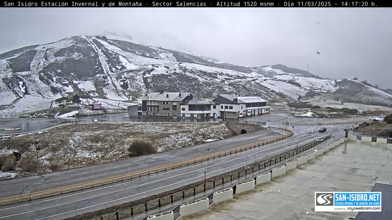 A snow-dusted mountainside with ski slopes overlooks a road and buildings in overcast conditions.