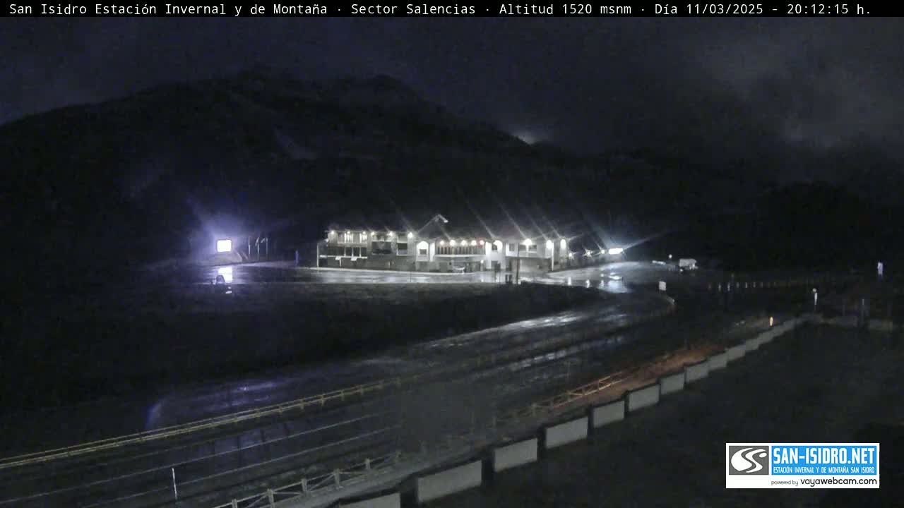 A nighttime view of a building complex illuminated by lights, situated near a mountain under a dark, possibly rainy sky.