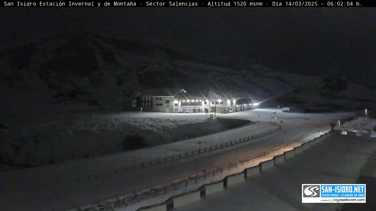 A snow-covered mountainside at night, illuminated by lights from a building near a roadway.