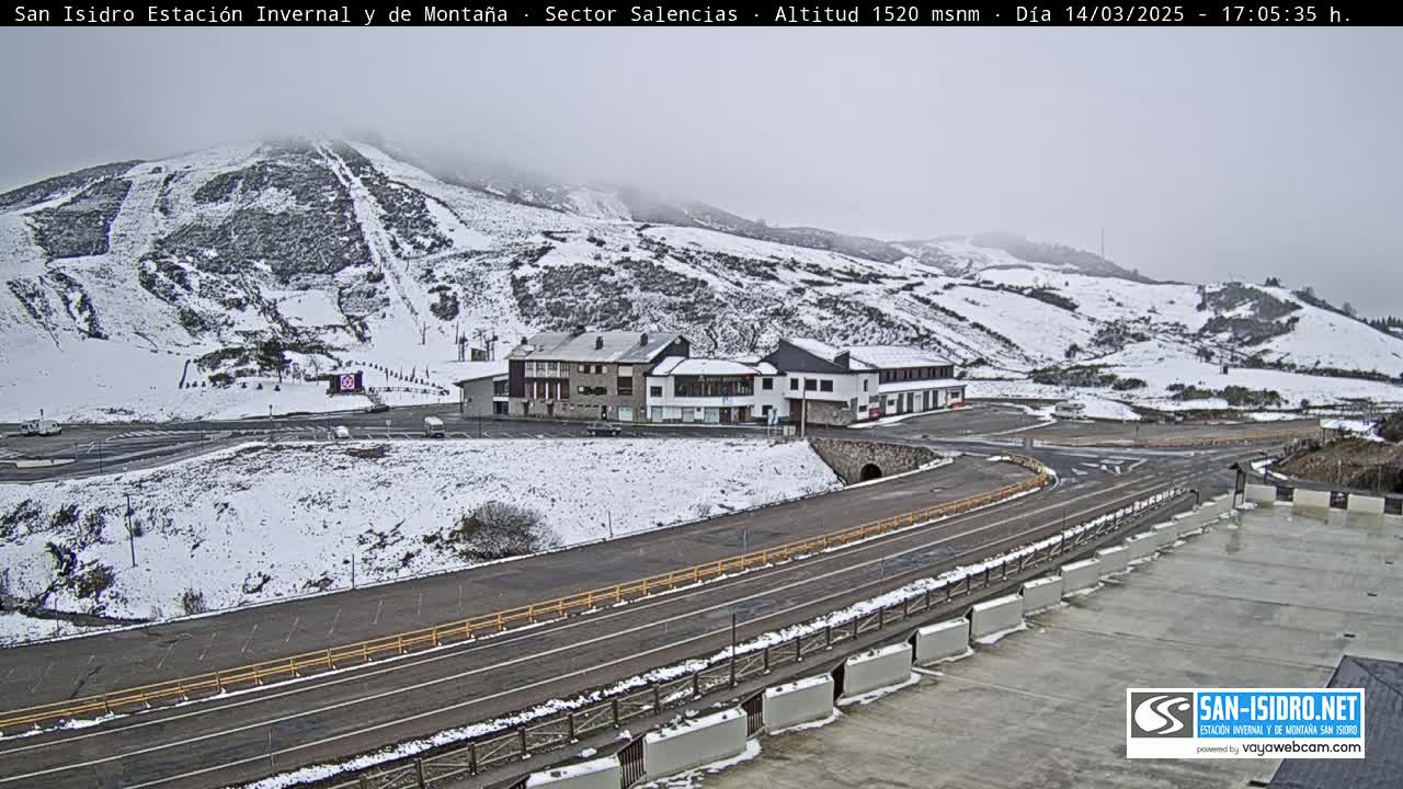 A snow-covered mountainside with ski slopes, a cluster of buildings, and a paved road are visible in overcast conditions.