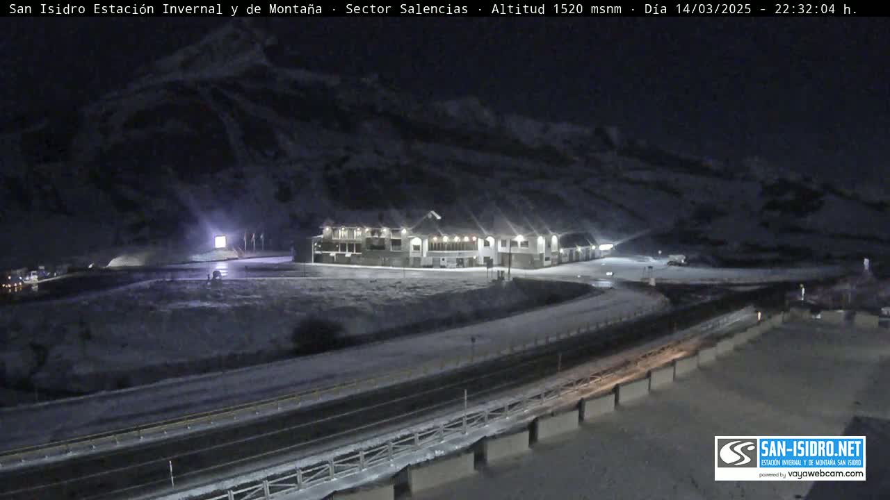 A snow-covered mountainside at night, with a building and a roadway visible below.