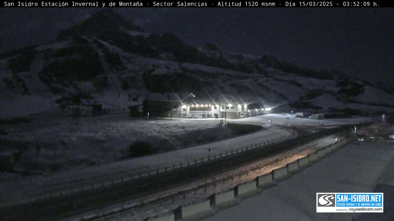 A snow-covered mountainside at night with a building illuminated, a road, and guardrails.