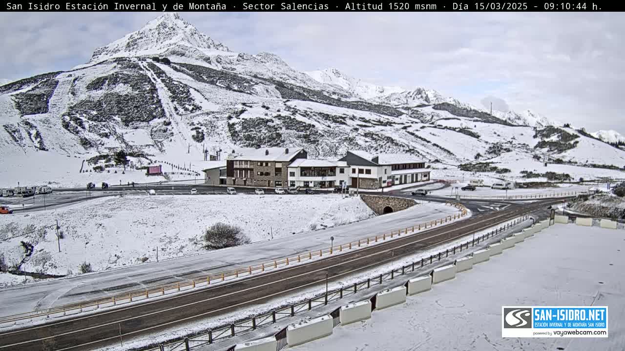 A snow-covered mountainside with ski slopes, a building complex, and a roadway are visible under an overcast sky.