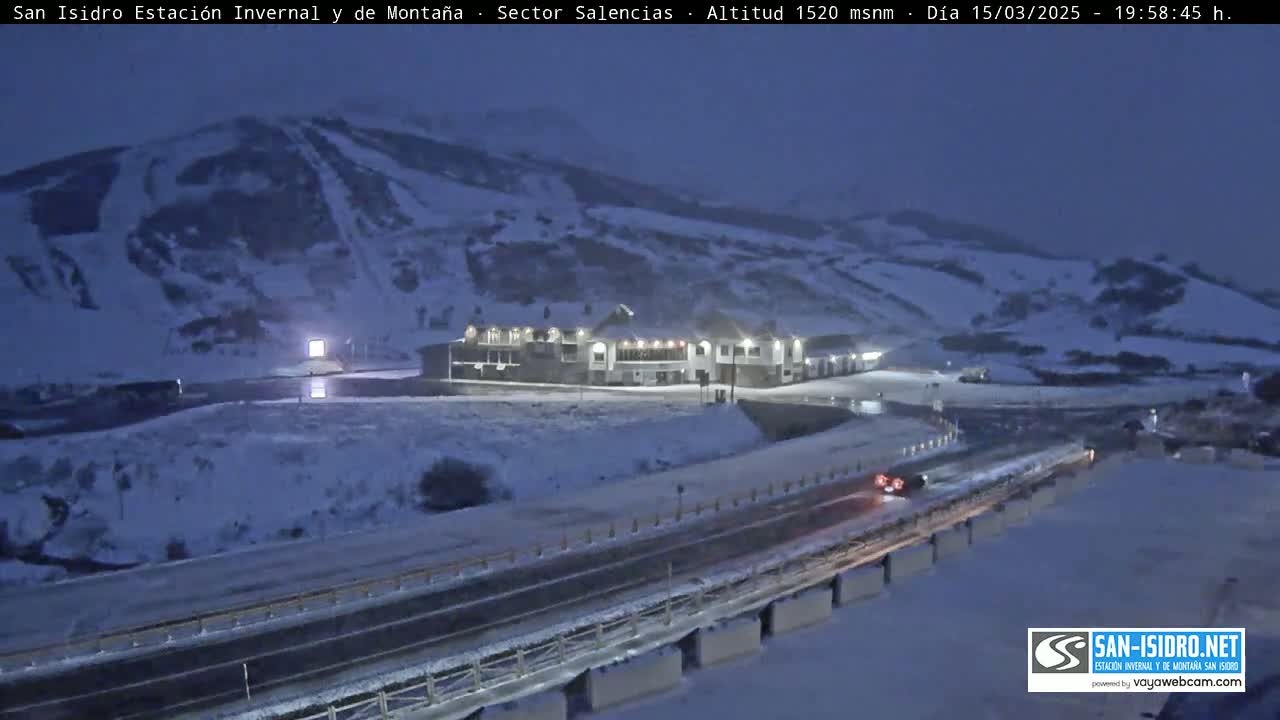 A snow-covered mountain slope with ski runs, a lit building, and a road with a vehicle are visible at night under a dark sky.