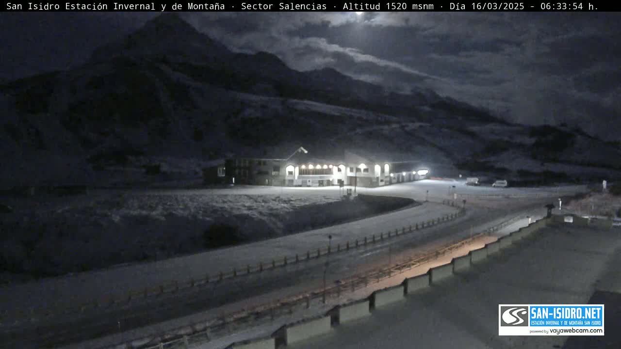 A snow-covered mountainside at night, with a lit building and roadway visible under a partly cloudy sky.