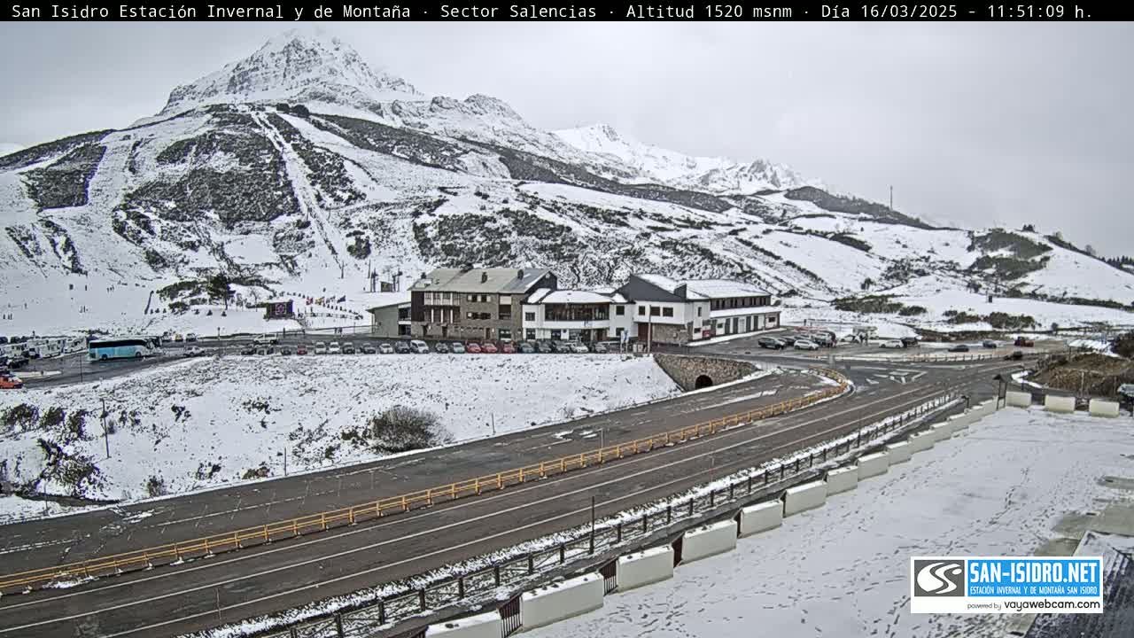 A snow-covered mountainside with ski slopes, a parking lot with vehicles, and a road winding through the scene under an overcast sky.