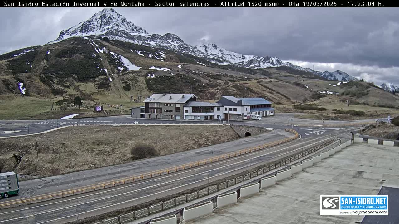 A mountain resort with snow-capped peaks under an overcast sky, featuring buildings, roads, and a large parking area.