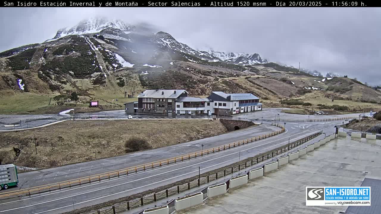 A mountain resort with two buildings sits beside a road curving through a parking lot, under an overcast sky with snow dusting the surrounding mountains.