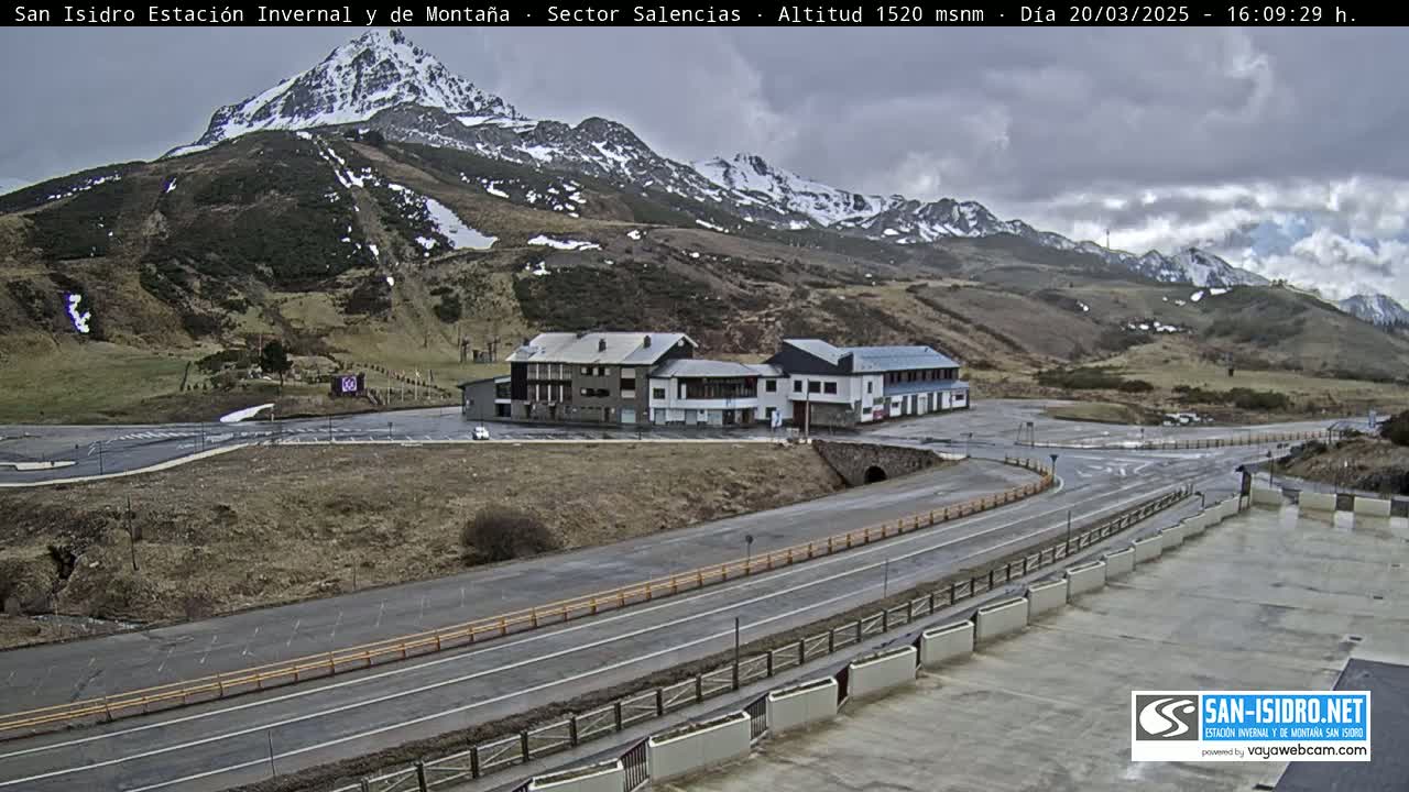 A mountain resort area with a few buildings, parking lots, and roads under cloudy skies, with snow visible on the distant mountains.