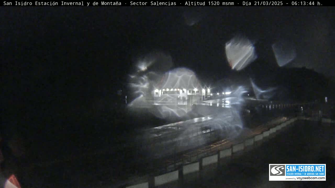 A nighttime, rain-swept scene shows a building and road, with streaks of light reflecting on a wet surface.