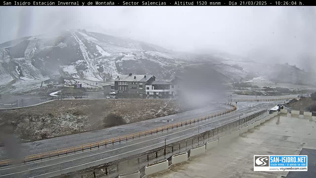 A snow-covered mountain with ski slopes is visible in the distance, alongside a wet road and buildings, under overcast skies.