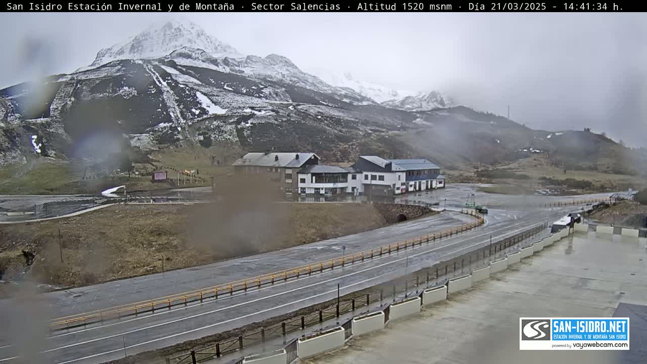 A wet, overcast day reveals a mountain road curving past a cluster of buildings nestled at the base of a snow-dusted mountain.