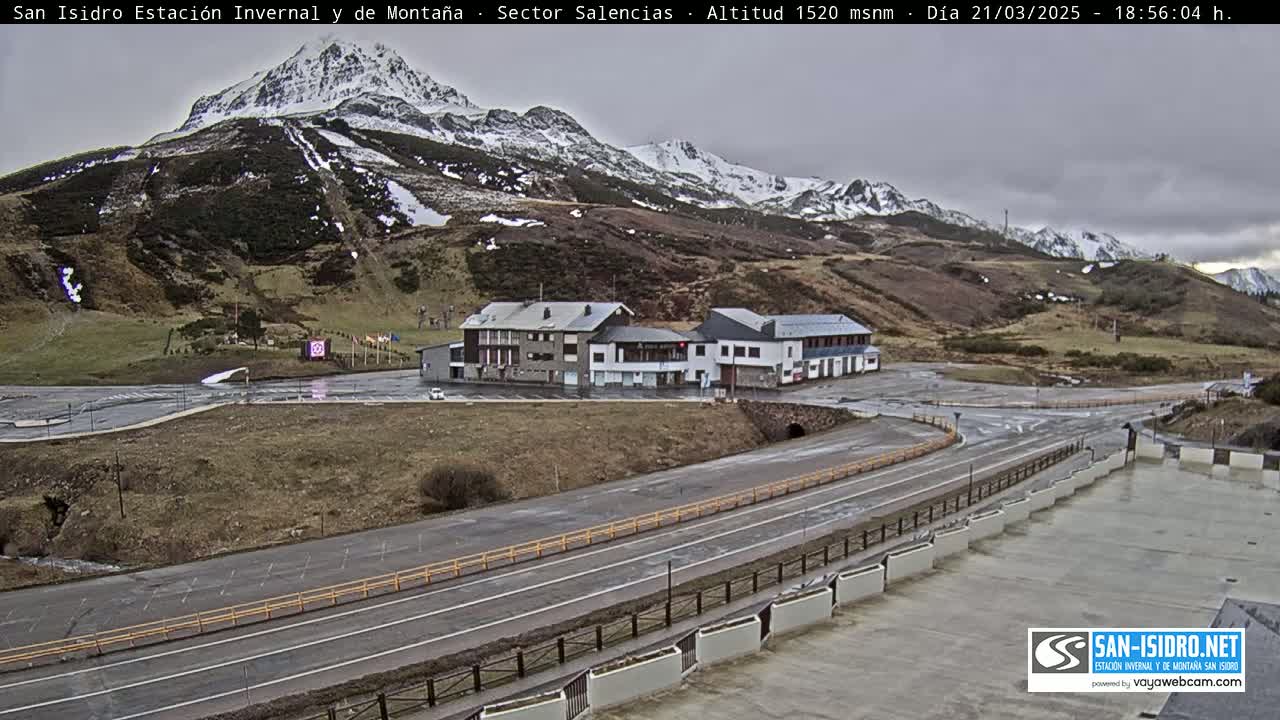 A mountainside ski resort with snow-capped peaks under an overcast sky, featuring a parking lot and several buildings.