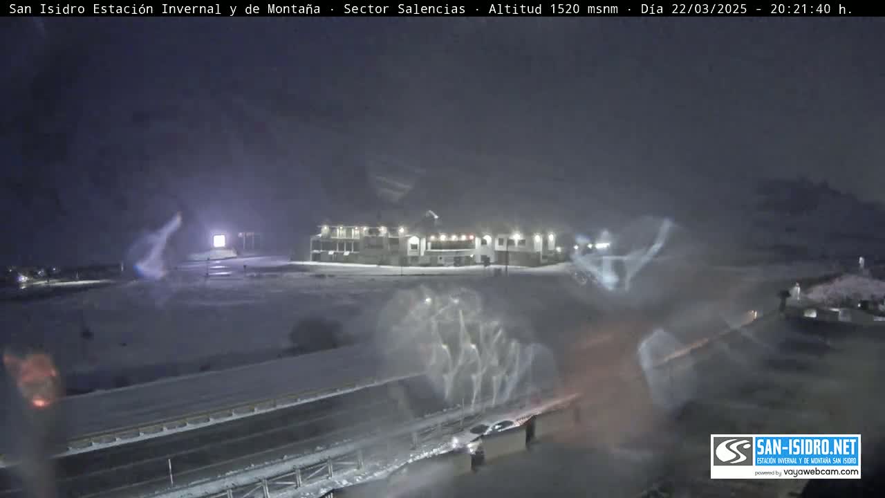 A snow-covered landscape at night, featuring a building and a highway with some vehicles, under a dark, snowy sky.