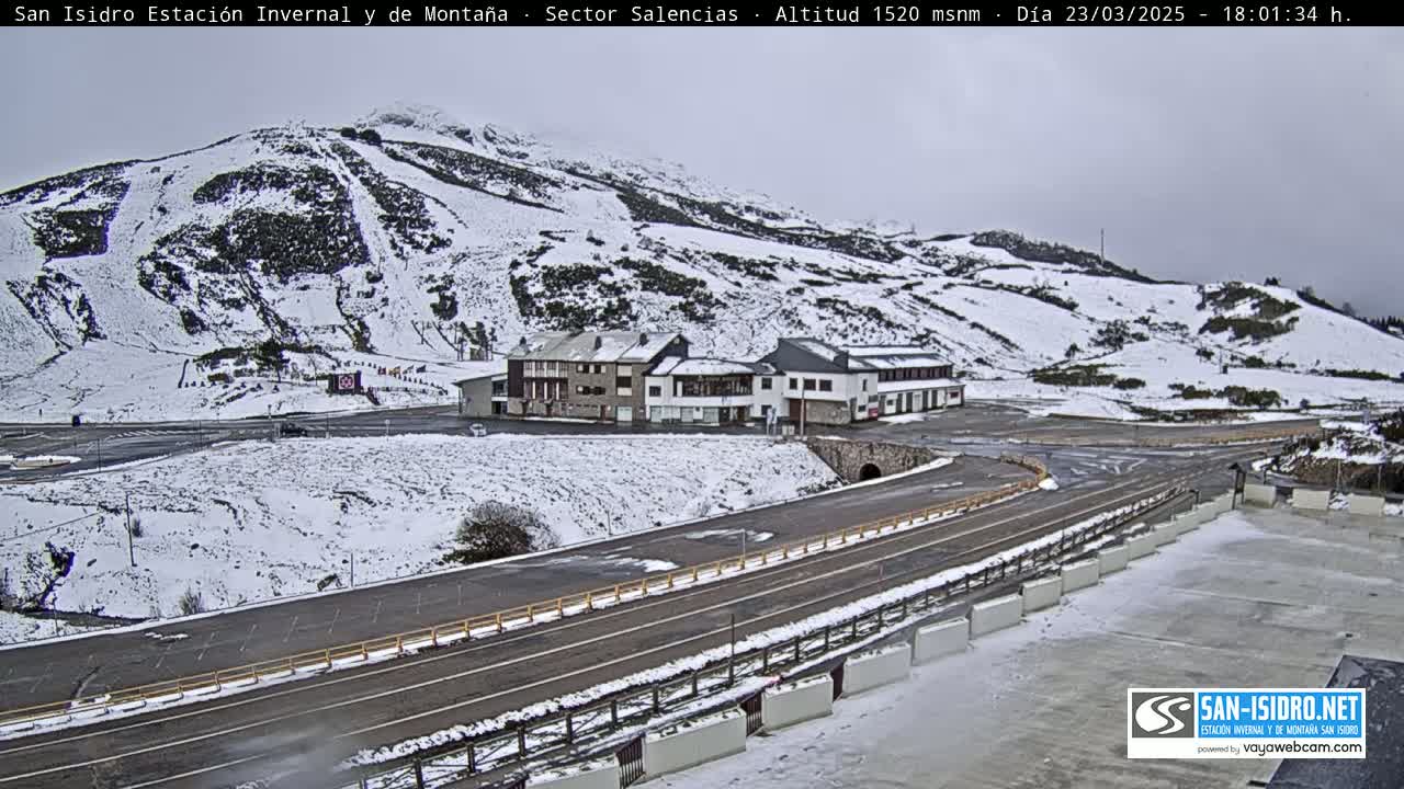 A snow-covered mountainside with a road and buildings, under an overcast sky.