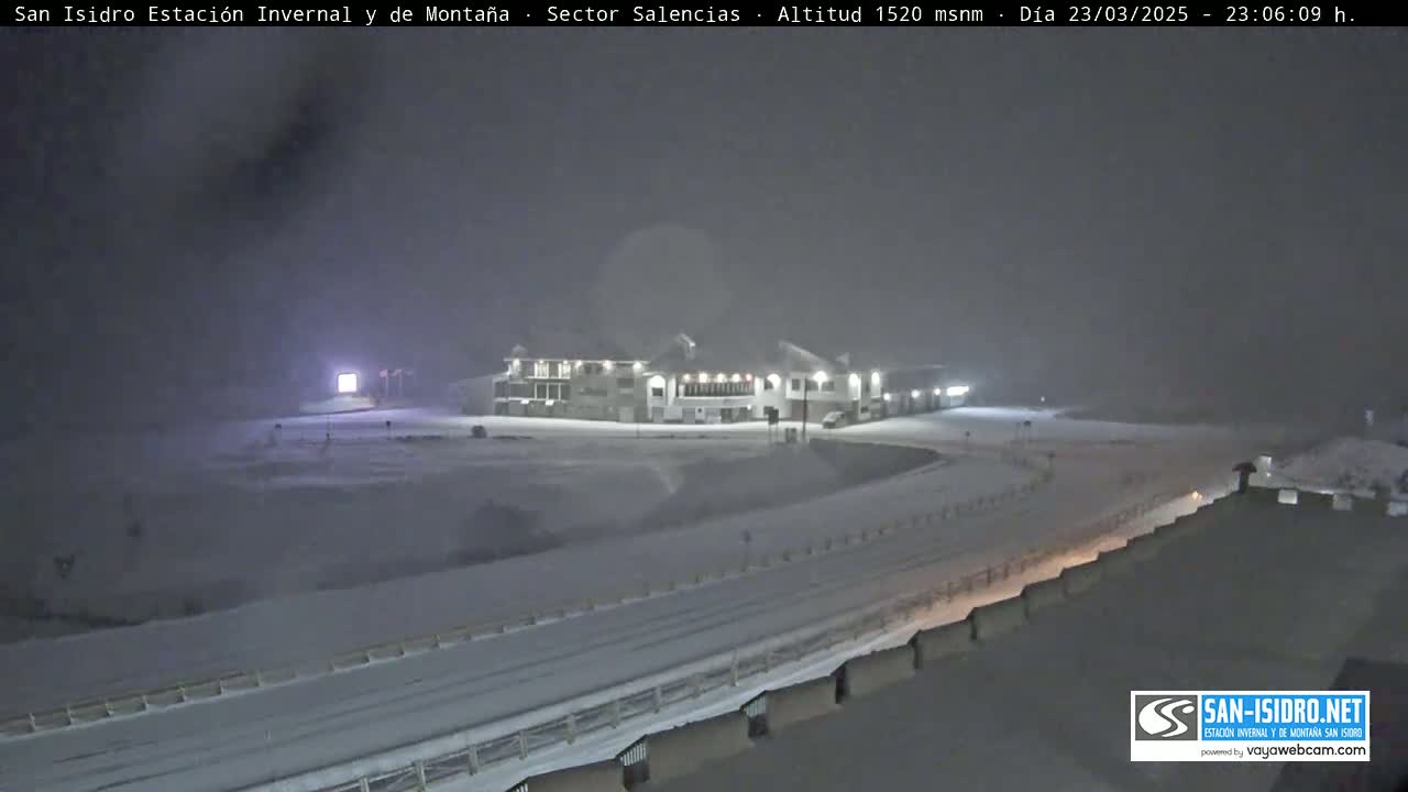 A snow-covered landscape at night features a large building illuminated by lights, with a road and railings visible in the foreground under heavy snowfall.