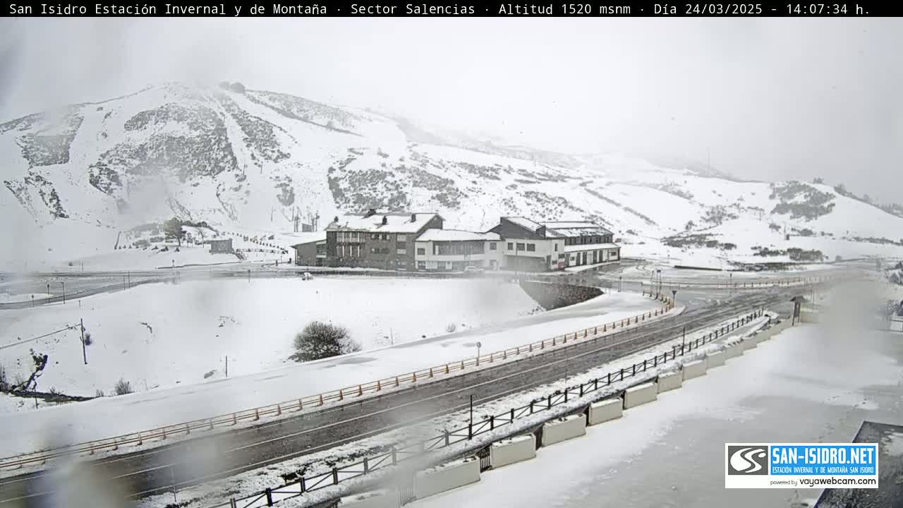 A snow-covered mountain slope with ski runs, a road, and a building complex is seen in snowy conditions.