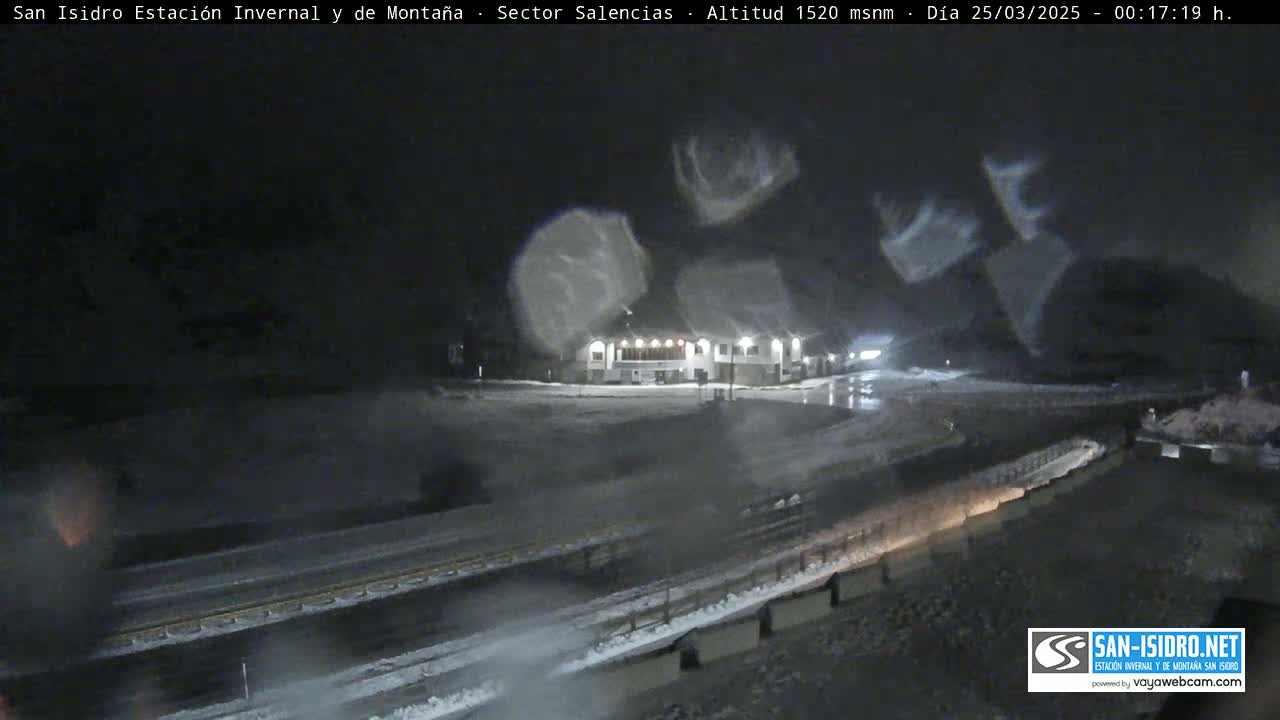 A nighttime, snowy scene shows a building and roadway with blurry snow falling.