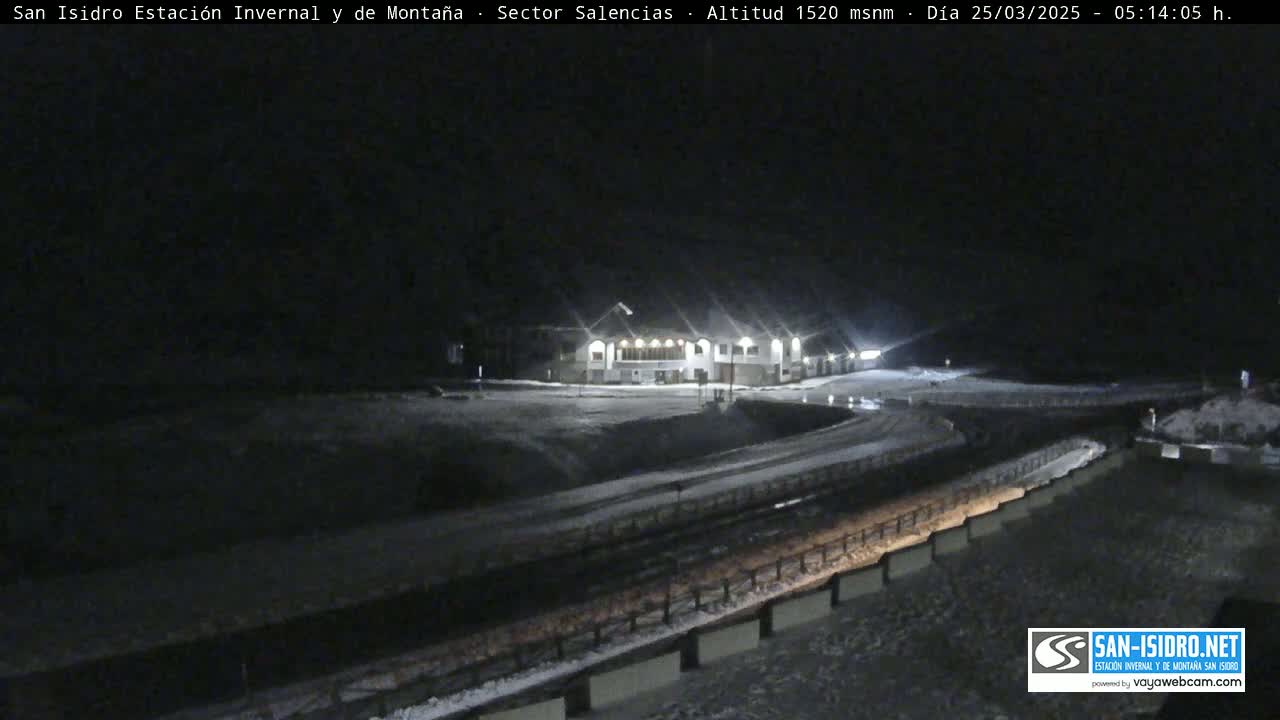 A snow-covered nighttime scene shows a building lit up, a roadway curving away from it, and a low wall along the road.