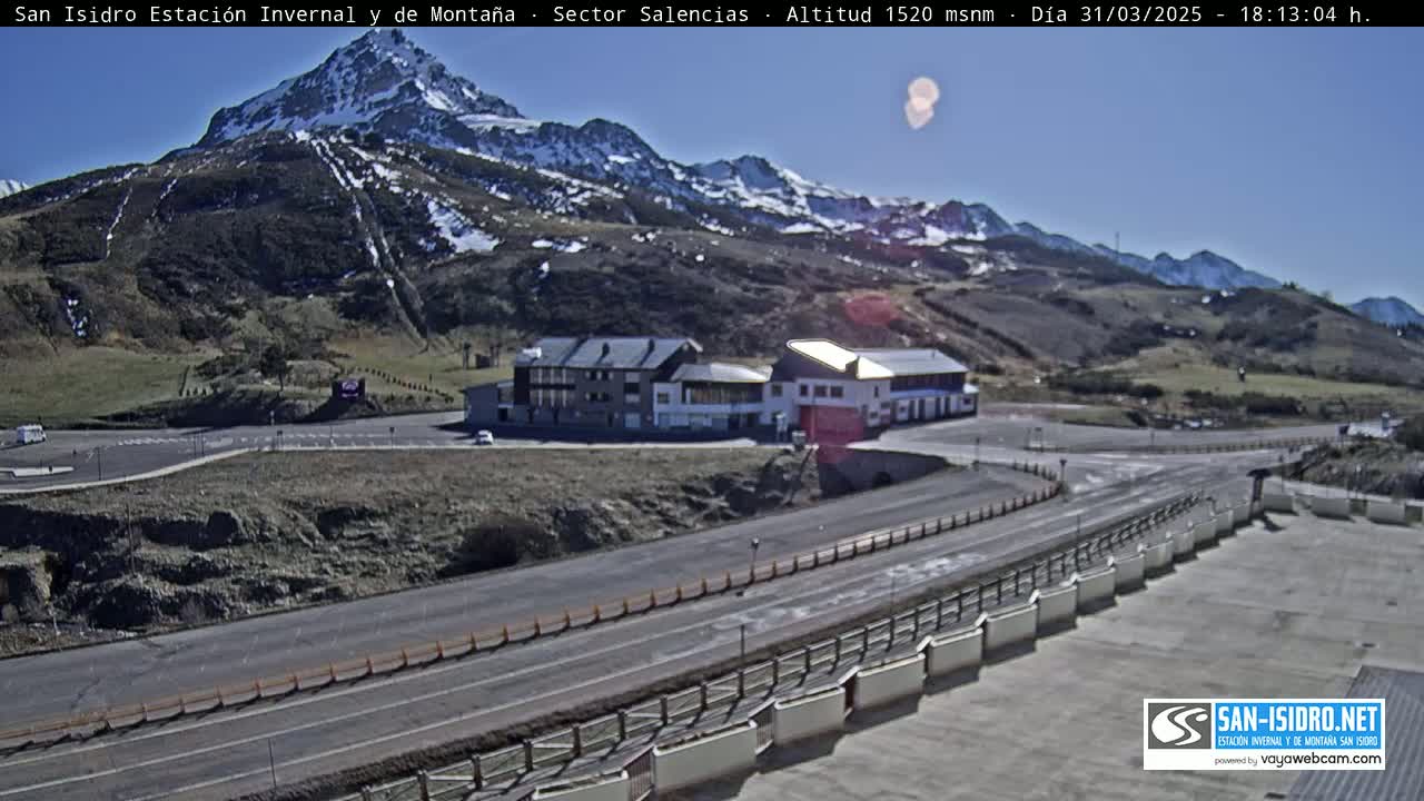 A mountainside resort with a parking lot and roads, viewed under a mostly clear, sunny sky with snow-capped mountains in the background.