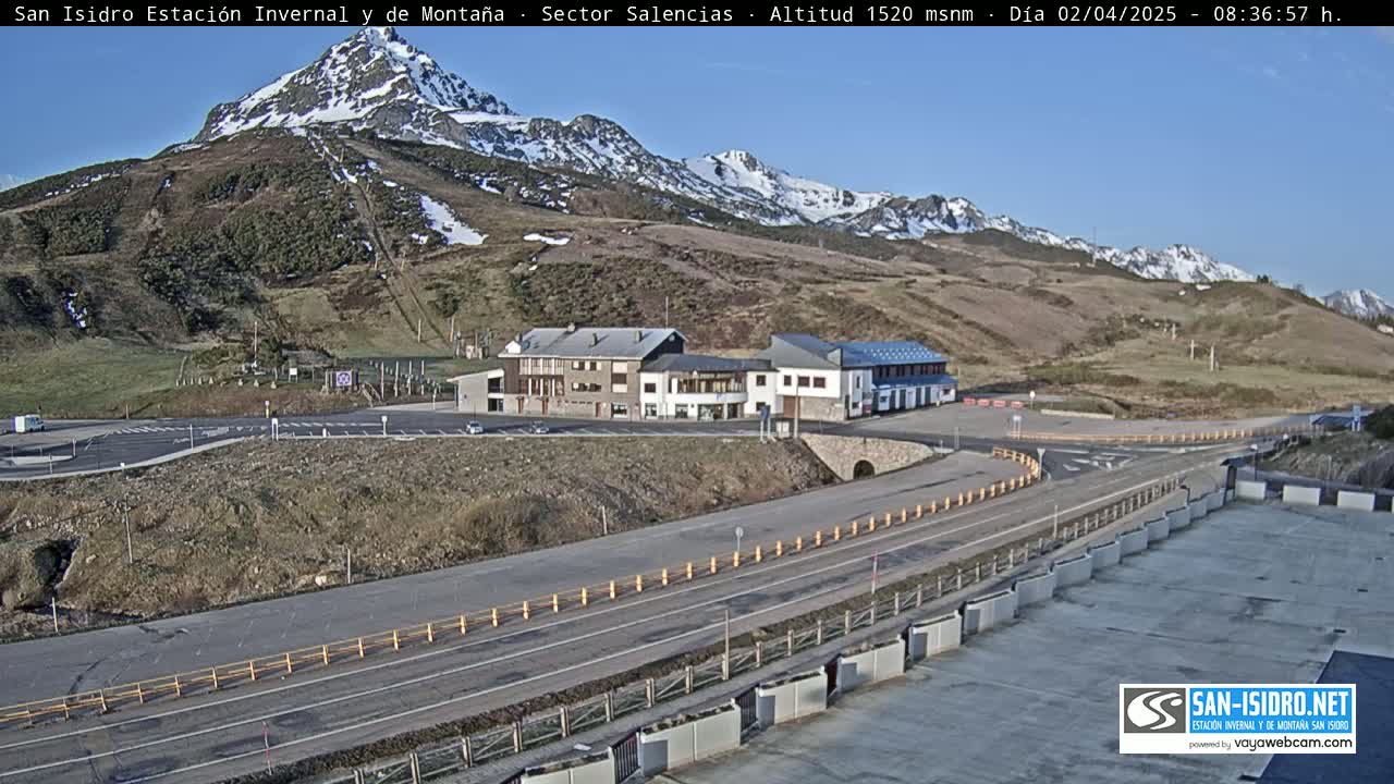 A mountain road winds past a cluster of buildings near a ski resort, with snow-capped mountains in the background under a clear, sunny sky.