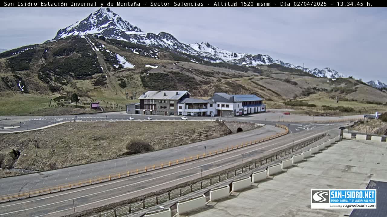 A mountain resort with a parking lot and several buildings sits below a snow-capped mountain range on a partly cloudy day.