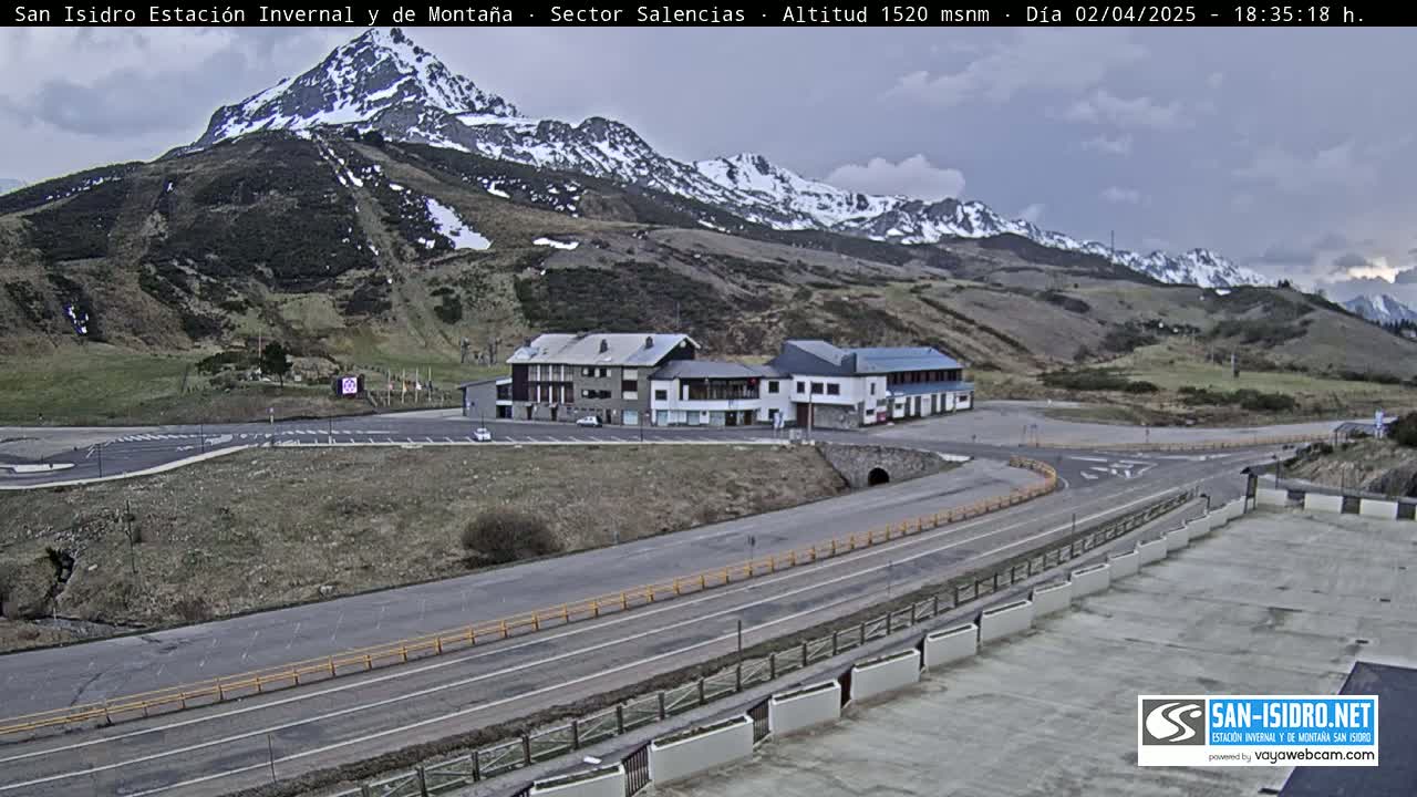 A mountain road curves past a cluster of buildings near a snow-dusted mountain range under an overcast sky.